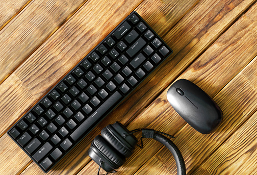 Small compact black computer keyboard next to a computer mouse and headphones on a table made of wooden pine boards. Wireless peripherals for gadgets and computers. Copy space. Daylight