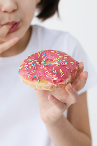 Little girl is eating pink doughnut with sprinkles in front of white background.