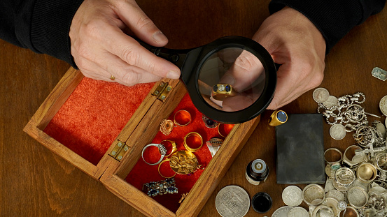 the jeweler, through magnifying glass, examines a ring with a diamond, against the background of a brown table on which precious things lie: rings, pendants, coins made of gold and silver.