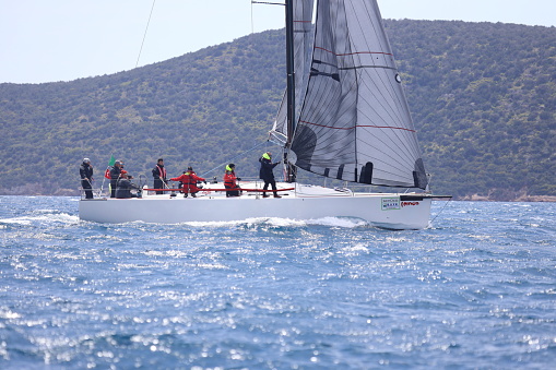 Bodrum,Mugla, Turkey. April 16,  2023: sailor team driving sail boat in motion, sailboat wheeling with water splashes, mountains and seascape on background. Sailboats sail in windy weather in the blue waters of the Aegean Sea, on the shores of the famous holiday destination Bodrum