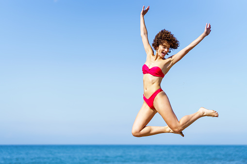 Smiling carefree young female with curly red hair in pink bikini, raising arms and jumping with excitement in air while looking down in daylight against blurred sea and blue sky