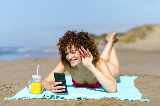 Young happy female with ginger curly hair lying on sandy shore with orange juice and waving hand while having video call on phone