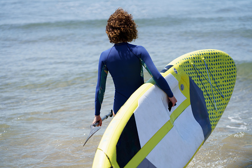 Back view of unrecognizable female surfer in wetsuit with SUP board and paddle standing in water at beach in rippling blue sea at daylight