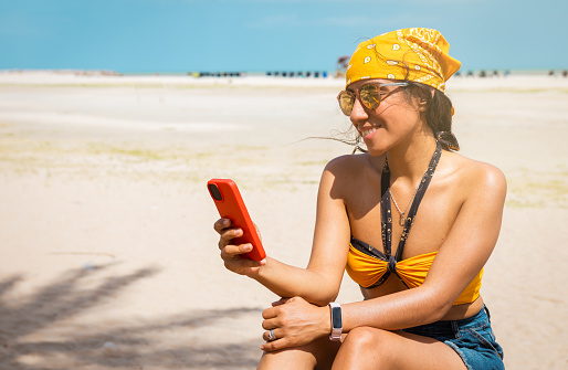 A beaming Latina woman with sunglasses and a headscarf captures a moment of pure joy on a sunny beach, phone in hand, as she basks in the warmth of the sun