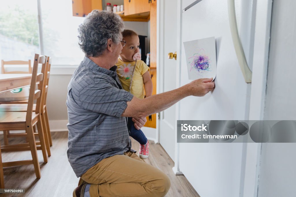 Grandfather with Granddaughter Putting Up Pre School Artwork on Refrigerator A proud grandfather with his 2 year old granddaughter putting up some beautiful artwork she made in pre School. Refrigerator Stock Photo
