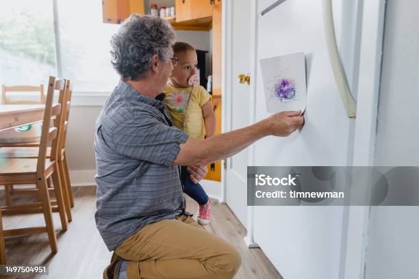 Grandfather With Granddaughter Putting Up Pre School Artwork On Refrigerator Stock Photo - Download Image Now