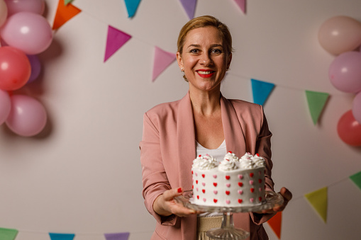Copy space shot of charming mid adult woman standing against a white background with colorful bunting garland and balloons on the wall, holding a birthday cake decorated with love hearts. She is looking at camera and smiling joyfully.