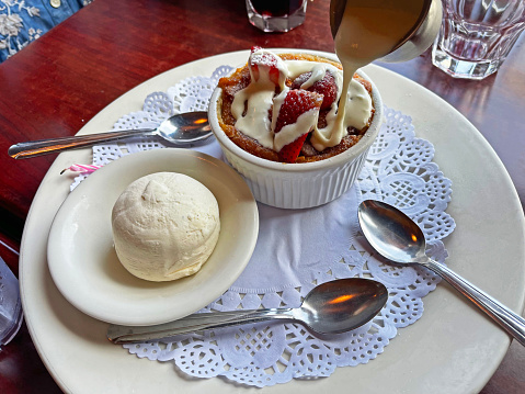 Bread pudding with cream and strawberries.   Side bowl if vanilla ice cream.   Three silver spoons.   All on doily and a large, white plate.  Cream pouring onto bread pudding.