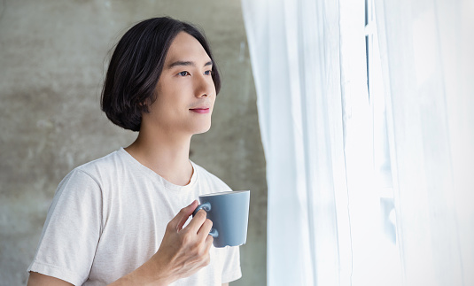 Portrait of young handsome asian man hand on coffee mug in bedroom. Smile happy asian boy in bed morning lifestyle near window. Japanese man leisure time on weekend, minimal modern break time concept