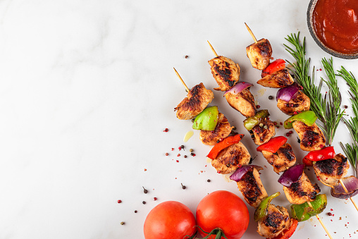 Grilled barbecue meat on skewers with vegetables, spices, basil and tomato ketchup on a plate over white background. Top view with copy space