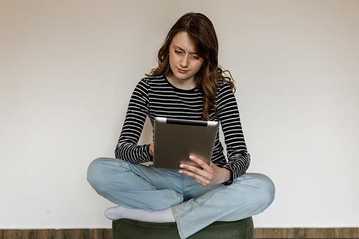 Full length shot of charming young woman sitting on a cozy, little green stool and using digital tablet.