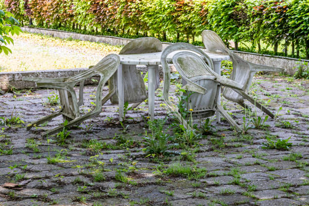Old dirty old plastic chairs and table on a back garden terrace, dirt, weathered and covered with mold, unkempt floor with wild plants, sunny spring day, green bush in the background