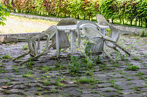 Old dirty old plastic chairs and table on a back garden terrace, dirt, weathered and covered with mold, unkempt floor with wild plants, sunny spring day, green bush in the background