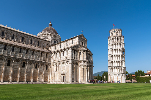 the Leaning Tower of Pisa in all its glory. Majestically, the tower rises into the sky and fascinates with its unique inclination. The bright blue sky forms a breathtaking backdrop for the imposing structure. The warm sunlight casts soft shadows on the marble and emphasizes the fine architectural details. The tower is surrounded by a picturesque landscape with green meadows and historic buildings, which give the photo an idyllic atmosphere.