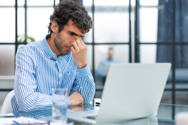 istock Handsome young business man with closed eyes touching face with hands while sitting on working place in office. Collegue is on the background. 1497613675