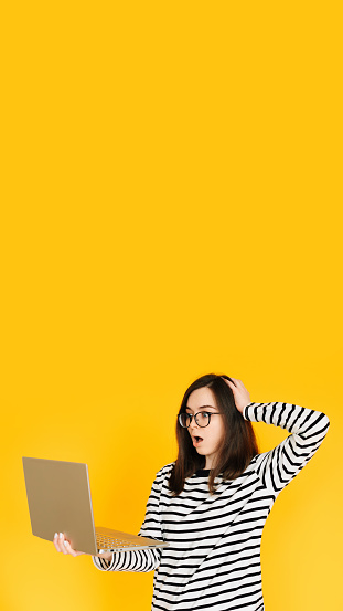 Picture of a cute impressionable lady in a striped T-shirt with glasses, reading the news or writing on a modern laptop device, hand head isolated yellow background color.