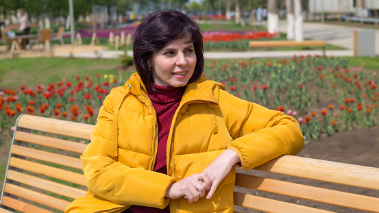 An adult happy woman with a smile face in a bright yellow jacket is resting on a bench in the park.