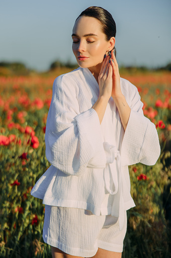 Young woman in white clothes walking on meadow and enjoying by flowering poppies with closed eyes at sunset.