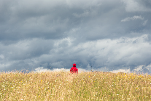 A lonely man in a red hoodie, standing atop a high grass mountain hill under a dramatic cloudy sky and enjoying tranquility and solitude.