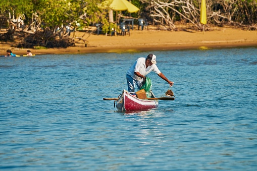Barra de Navidad, Mexico – February 19, 2022: A closeup of a  man in a small boat sailing on a tranquil lake  and a beach in the background
