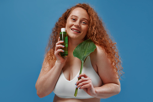Beautiful redhead plus size woman with freckles in white lingerie with bottle of lotion and leaf isolated on blue background. Body positive. Cosmetology and skin care.