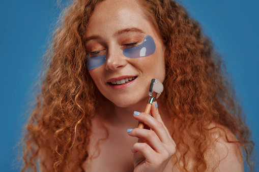 Women's plus size beauty, eye patch and face roller for studio dermatology, healthcare or skincare. A red-haired plump woman on a blue background. Body positive.