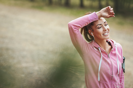 Happy woman, wearing sportswear, with earphones in her ears is listening music and resting in a break of exercises in a sunny forest.