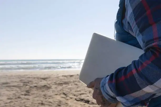 A young adult male standing on a beach with a laptop computer in his hand