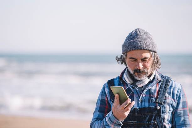 istock Elderly man wearing earphones while standing on the beach, using a mobile phone for communication 1497633970