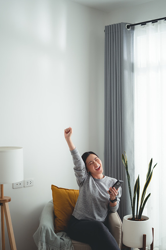 Asian woman holding and looking at smartphone happy excited and making a winning gesture sitting on armchair at home.