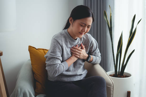Asian woman suffering from chest pain sitting on armchair in living room at home. Asian woman suffering from chest pain breathing problems holding hands on her chest sitting on sofa in living room at home. asian woman heart attack stock pictures, royalty-free photos & images