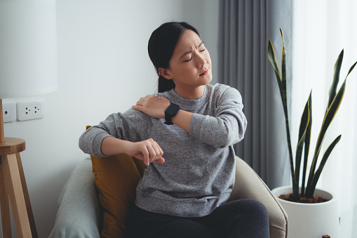Asian woman suffering from shoulder pain massaging on shoulder by hands sitting on armchair in living room at home.
