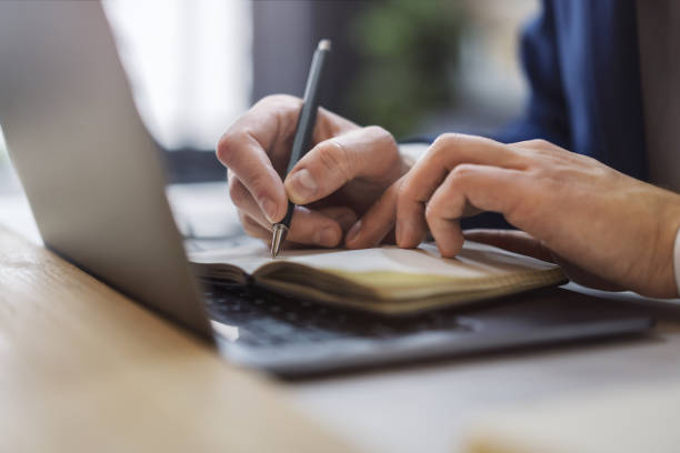istock Detailed view of a man's hand jotting down notes in a notepad on a modern laptop, with a blurred background 1497641911