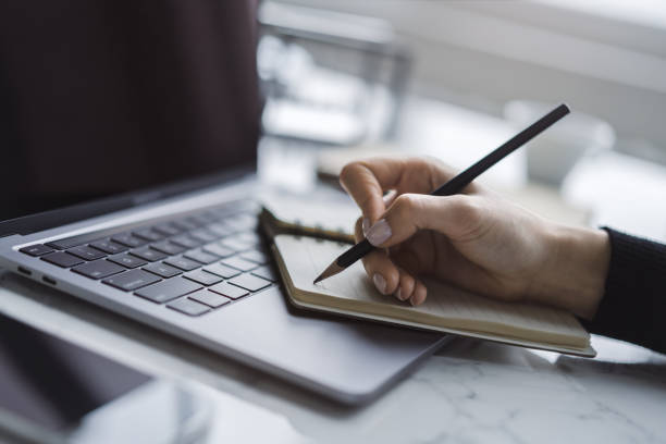 istock Tight focus on a woman's hand as she writes in a notepad on a contemporary laptop, background subtly blurred 1497642273