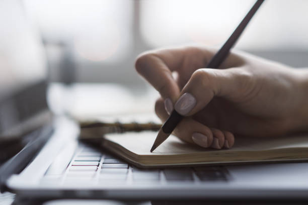 The image captures a woman's hand in close view as it writes in a notepad on a chic laptop, background out of focus