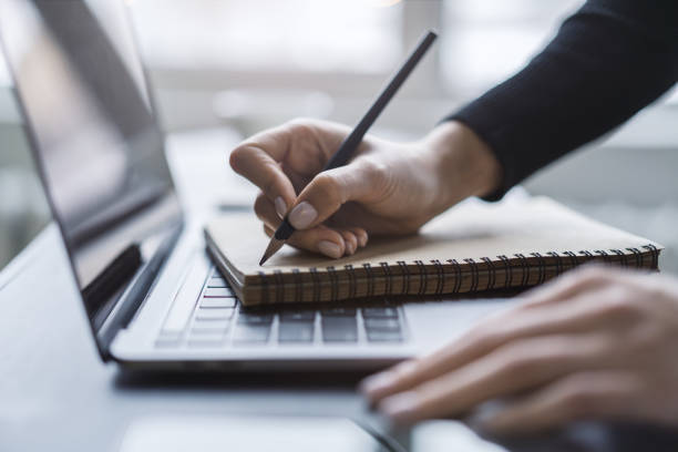 istock Detailed view of a woman's hand taking notes in a notepad on a cutting-edge laptop, against a blurred backdrop 1497643535