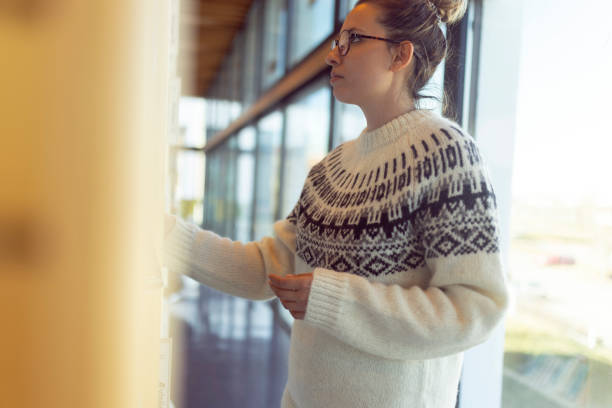 istock Side view of a concentrated female student in sweater reading book while standing next to bookshelf in library. Iceland 1497651781