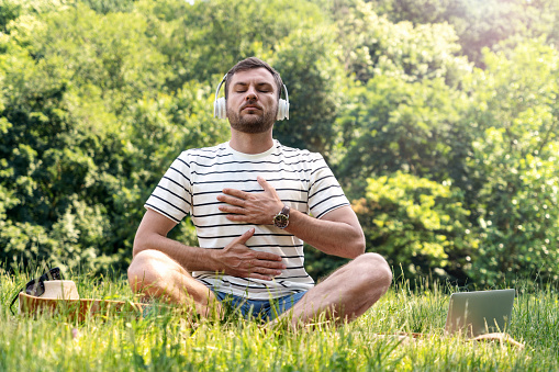 Man wearing headphones doing yoga breathing exercise in half lotus position in the park, listening music for meditation.