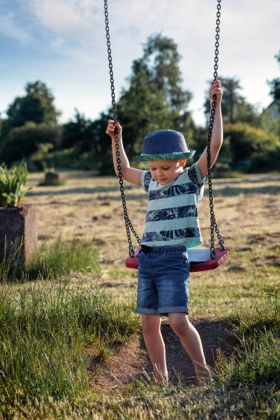 A little boy in a blue hat is trying to reach and climb the outdoor swing by himself stock photo