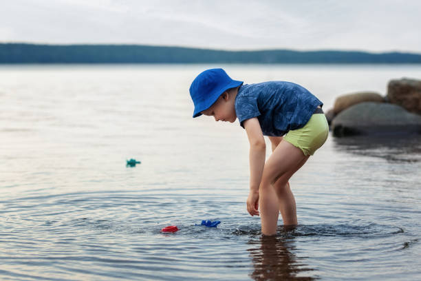 Young boy crouches down in shallow calm lake water and playing with his colored toy boats. It is summertime. stock photo