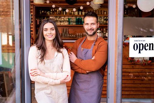 Small business owner woman and man standing at the cafÃ© door waiting for customers. Small business owners looking at camera and smiling.