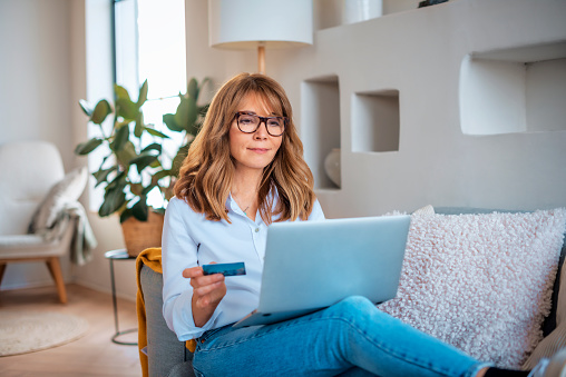 Mid aged woman sitting at home and using credit card and laptop for shopping online. Confident female wearing shirt and eyewear.