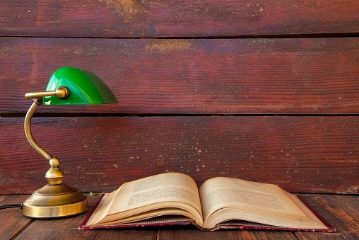 Vintage reading lamp. Antique copper-colored metal lamp with a green shade on a rustic wooden background next to an old reading book. Back to school concept.