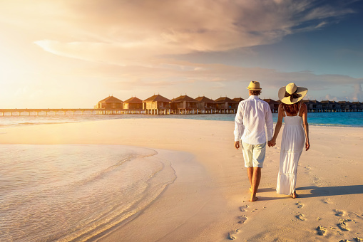 A beautiful traveler couple in white summer clothing walks along a tropical beach in the Maldives holding hands during sunset time
