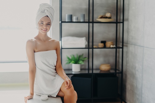Relaxed woman in bathroom, wrapped in towel, cares for skin with product. Spa and wellness concept.