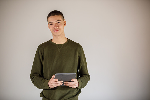 Copy space shot of confident teenage boy standing against white background, holding a digital tablet, looking at camera and smiling.