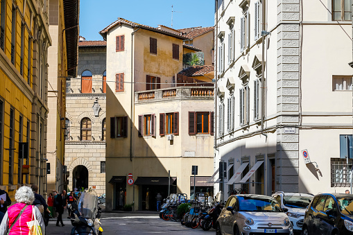 Florence, Italy - April 19, 2023: Street between buildings in the old town. There are parked cars, bicycles and scooters. People are walking on the sidewalk.