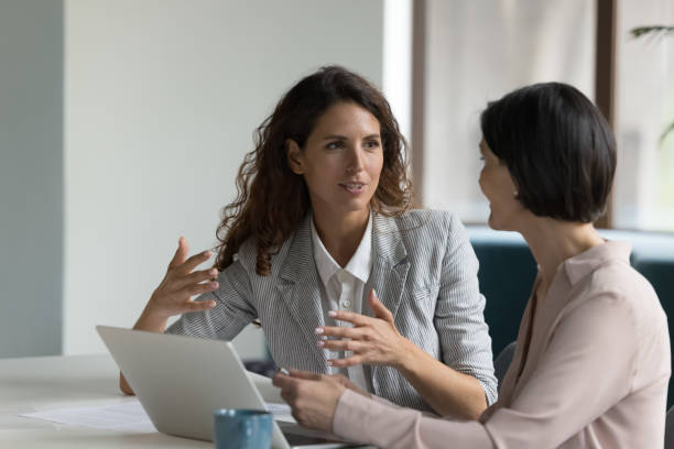 Two attractive businesswomen sit at desk discuss project details stock photo