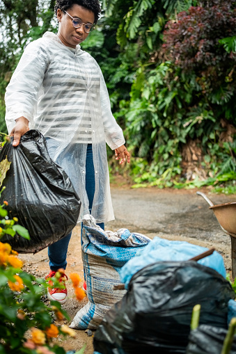 Young Brazilian woman wearing raincoat gathering garbage bags during garden cleanup in rain