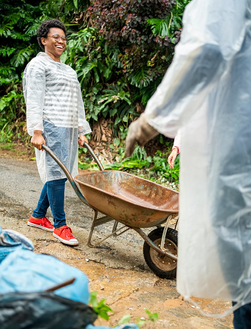 Smiling young african woman wearing raincoat holding a wheelbarrow walking in garden during cleanup work in a rainy day